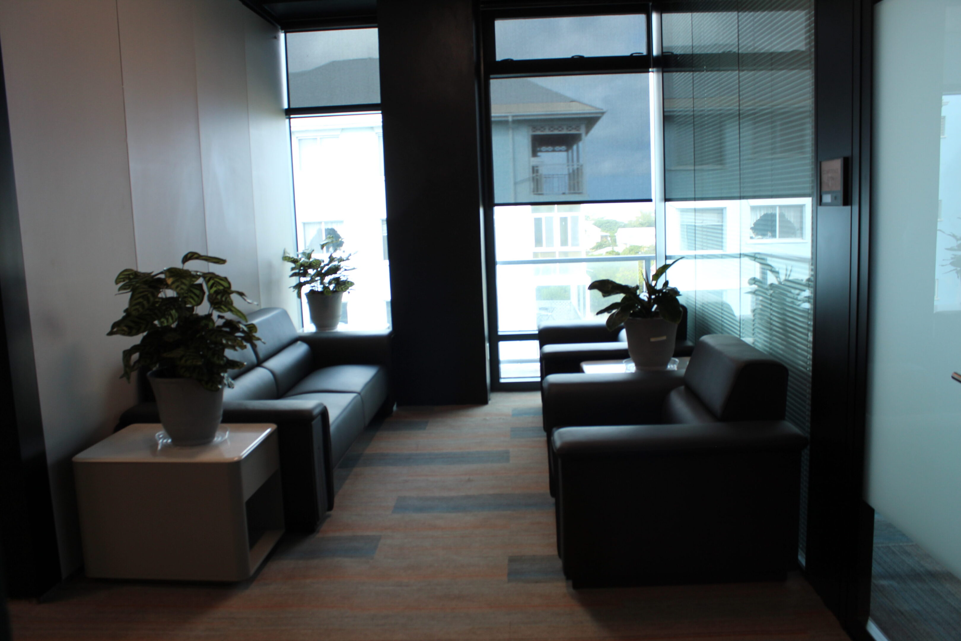 Modern office waiting area with black couches and potted plants near large windows.