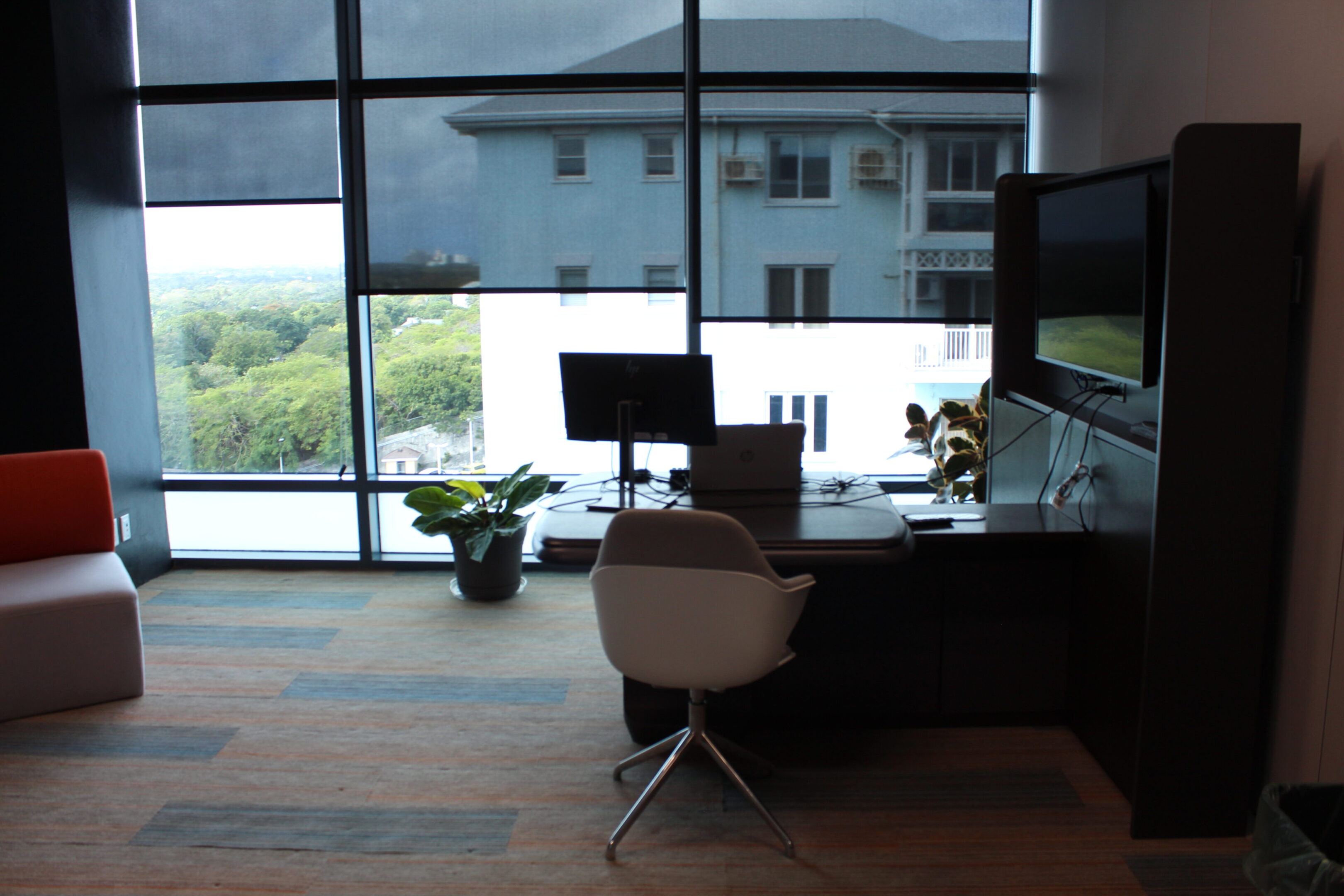 A modern office workspace with a chair, monitor, and potted plants near large windows.