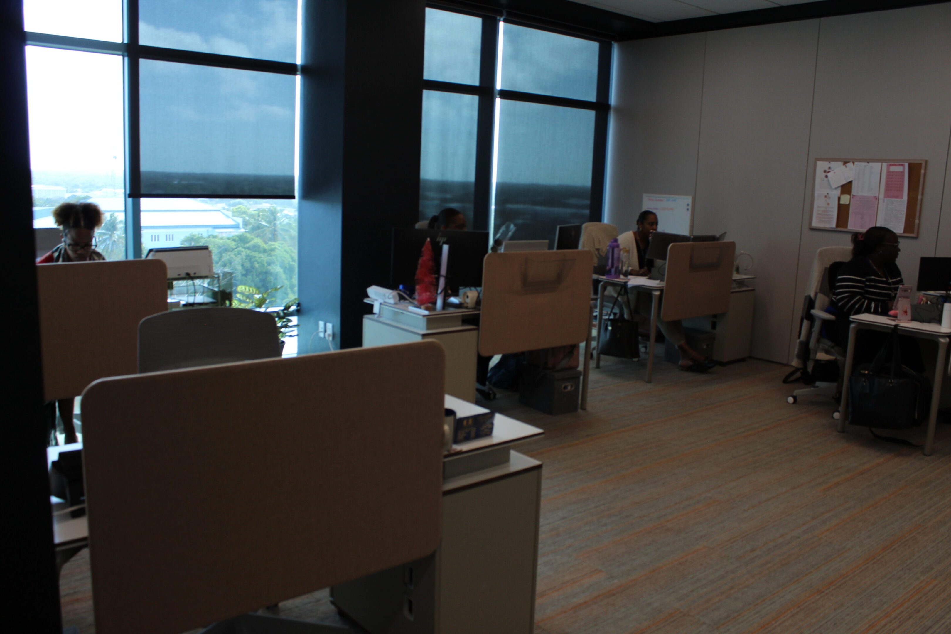 Office workspace with desks and chairs near large windows overlooking a cityscape.
