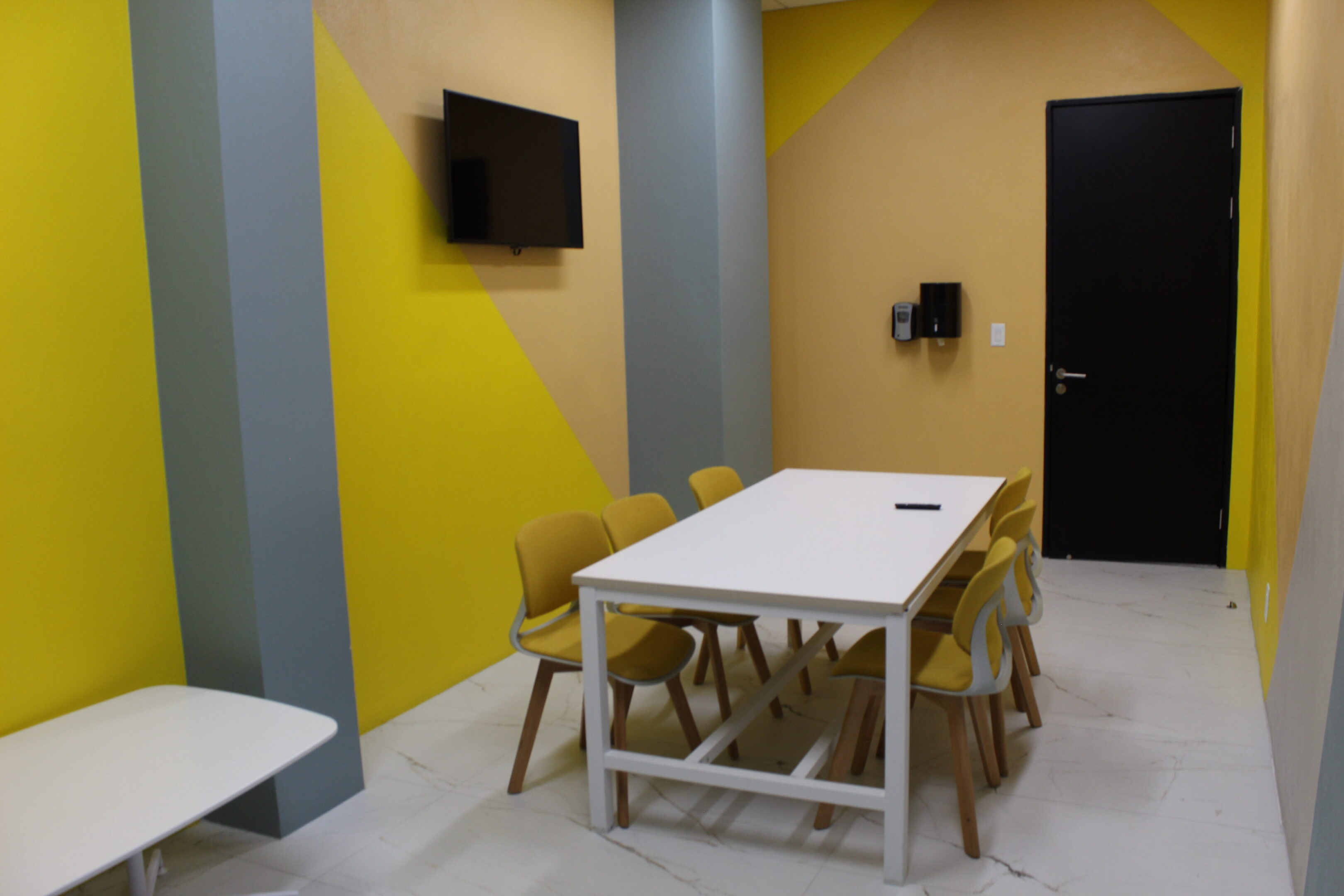 Modern meeting room with white table, yellow chairs, and wall-mounted TV.